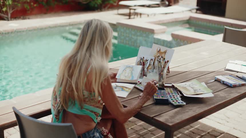 A creative woman with long blonde hair sits at a wooden table by a sunny pool, painting whimsical animal portraits with watercolors