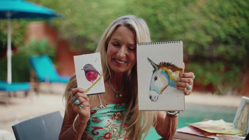 A smiling mature female artist with long blonde hair proudly presents her latest watercolor paintings of a bird and a donkey while sitting outdoors by a pool