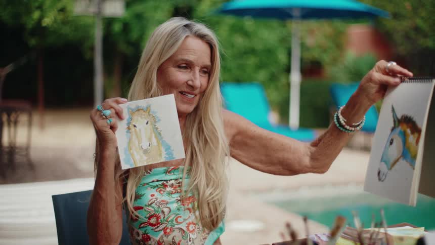 A happy senior woman proudly displays her watercolor paintings of a horse and a donkey by the pool