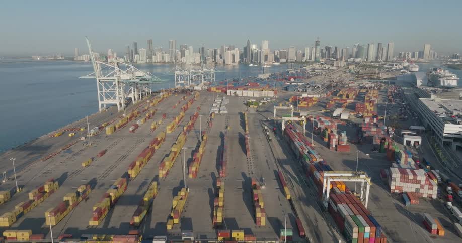 A slow aerial view moves over a large shipping container port with the Miami city skyline visible across the water at sunrise