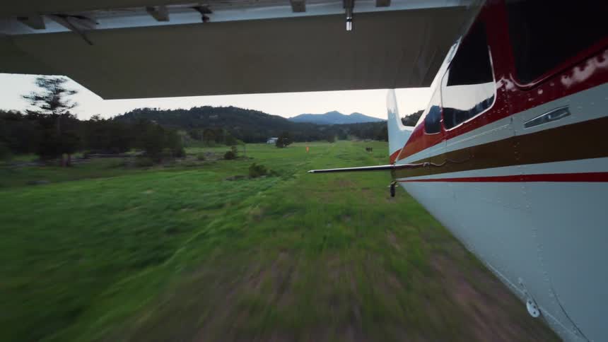 Point-of-view from a small airplane as it accelerates down a grassy runway and takes off into the sky above a forested mountain valley
