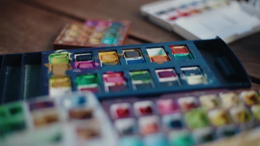 A slow, close-up shot pulls back to reveal multiple well-used watercolor paint palettes on a rustic wooden table