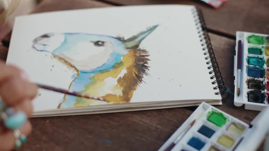An elderly artist with turquoise rings paints a colorful watercolor portrait of a donkey in a sketchbook
