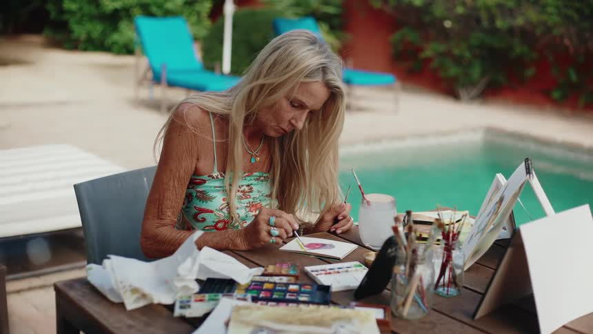 An artistic senior woman with long blonde hair concentrates on her watercolor painting at a table beside a swimming pool