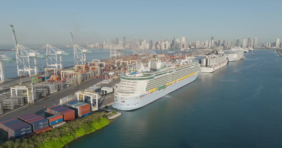 A slow aerial push-in reveals large cruise ships docked at a busy commercial shipping port with the downtown city skyline visible in the distance