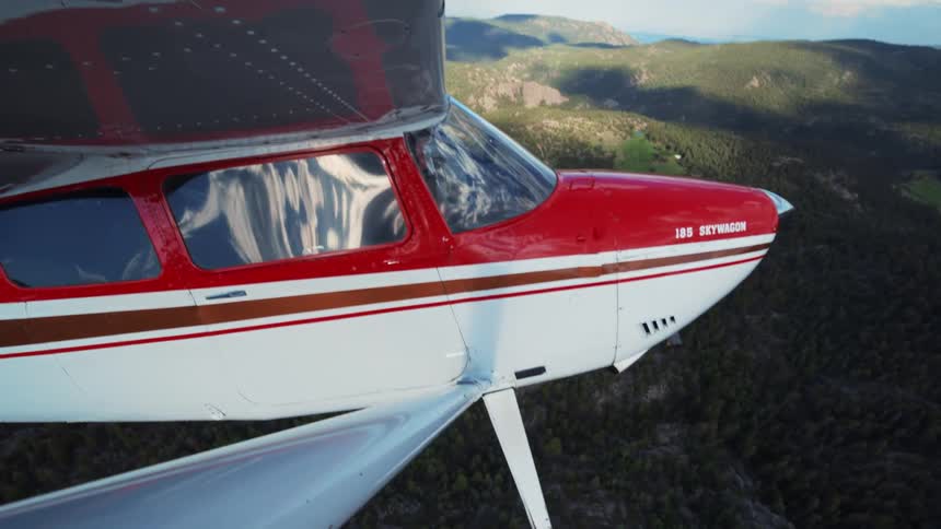 A pilot flies a red and white single-engine airplane over a vast, tree-covered mountain landscape at sunset