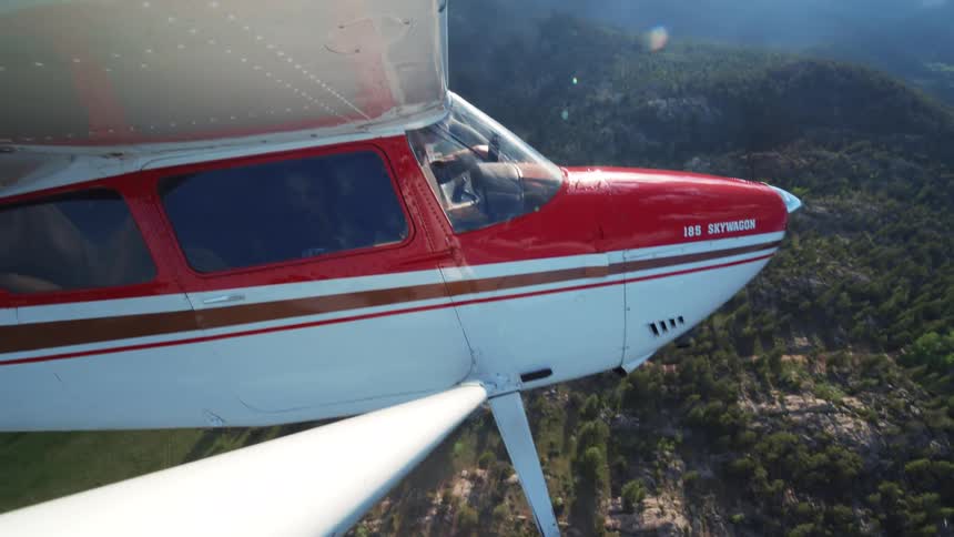 An aerial shot captures a small red and white propeller plane in flight over a vast, green mountain forest