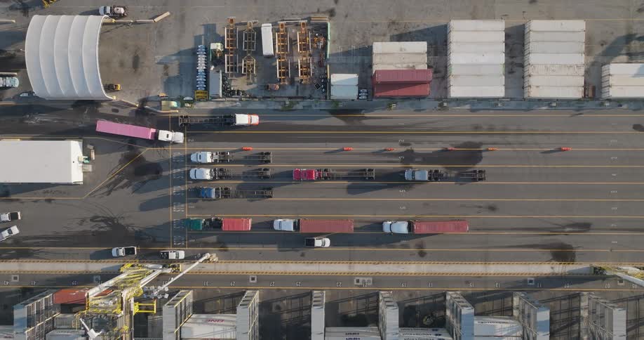 A static aerial top-down shot captures the organized movement of semi-trucks navigating lanes at a bustling industrial shipping port