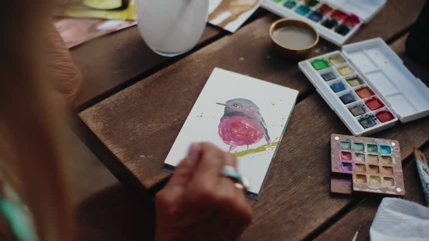 An over-the-shoulder view of an artist's hands painting a colorful bird with watercolors on a wooden table