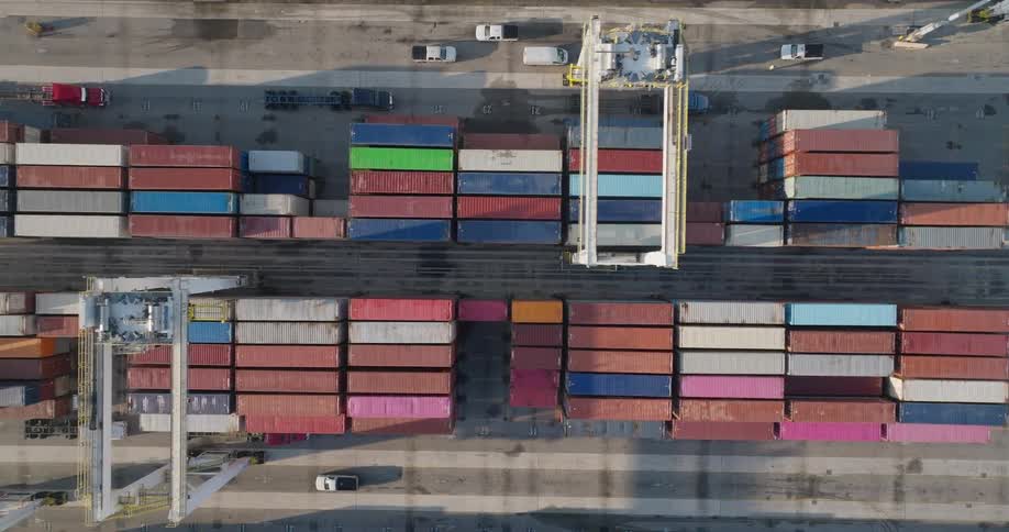 A gantry crane moves over stacks of colorful shipping containers at an industrial port in this top-down aerial shot