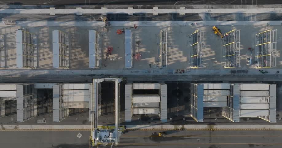 A top-down aerial view reveals a large gantry crane moving over stacked shipping containers at an industrial port under construction