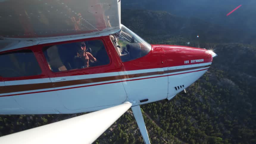 A woman takes a selfie from the window of a small Cessna airplane flying over a scenic, forested mountain range