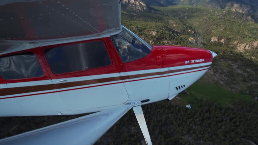A red and white 185 Skywagon propeller plane soars over a vast, tree-covered mountain range