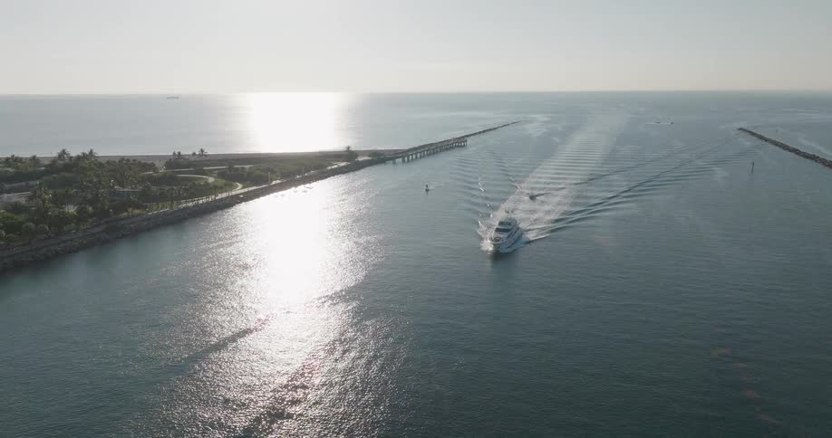 An aerial view follows a luxury yacht as it sails out of a coastal channel and into the open ocean on a bright, sunny day