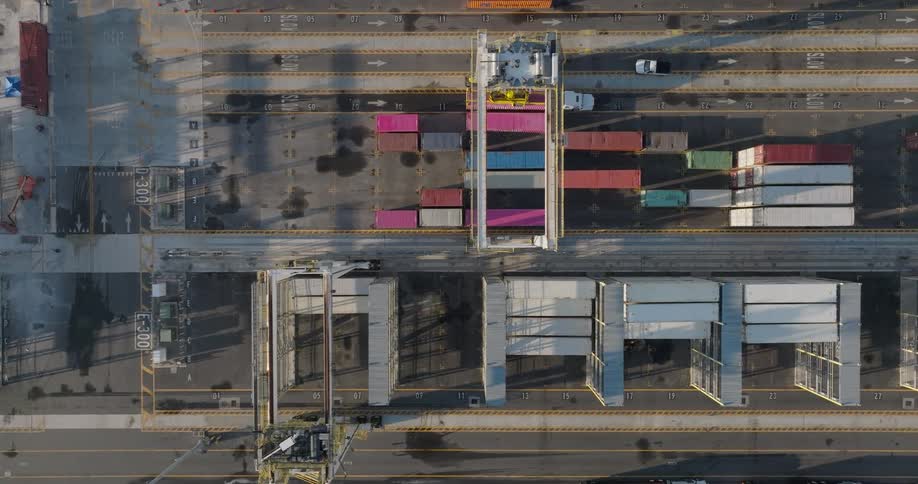 A top-down aerial view shows large gantry cranes moving along tracks and stacking colorful shipping containers at an industrial port terminal