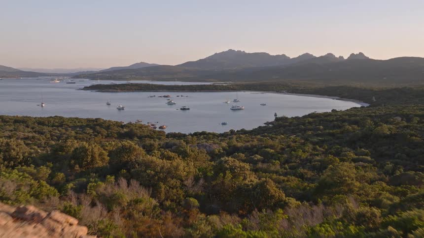 An aerial view flies over rocky, tree-covered hills to reveal a tranquil bay filled with anchored yachts at golden hour