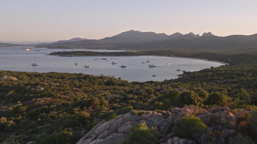 An aerial view flies over a rocky, green coastline to reveal a tranquil bay filled with sailboats and yachts at sunset