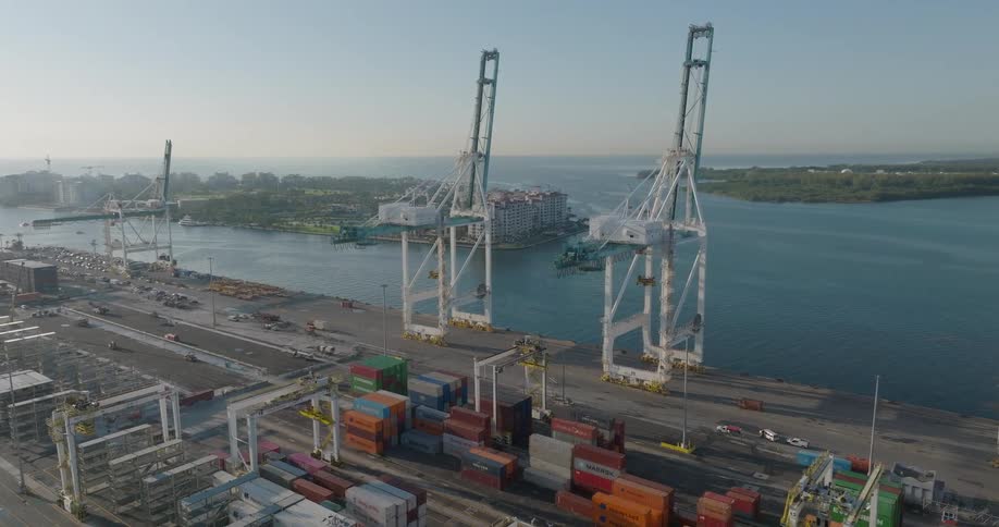An aerial view slowly pushes in on the massive gantry cranes and shipping containers at the bustling Port of Miami on a sunny day