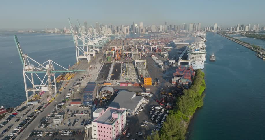 Slow aerial pullback revealing the bustling Port of Miami, featuring cranes, shipping containers, and a cruise ship with the city skyline in the distance
