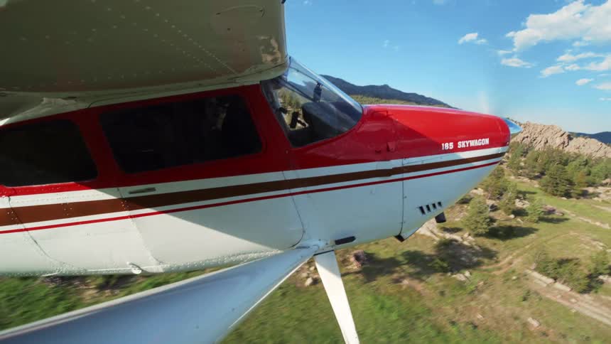 A small red and white propeller plane takes off from a grassy runway and flies over a scenic, forested mountain landscape