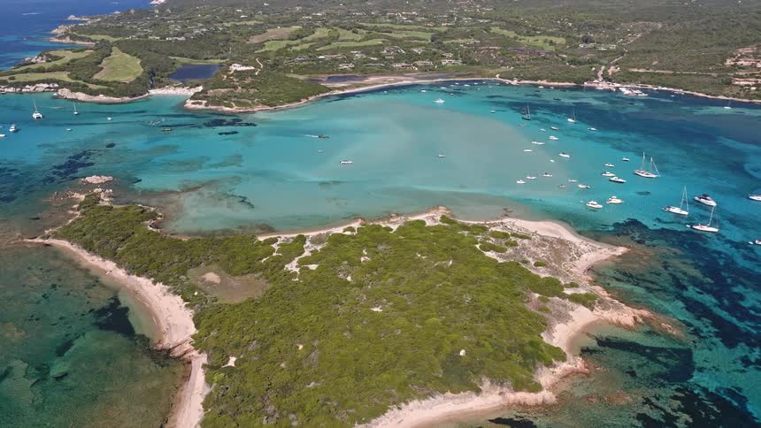 An incredible aerial view slowly rises over a lush green island surrounded by crystal-clear turquoise water in a bay filled with anchored yachts