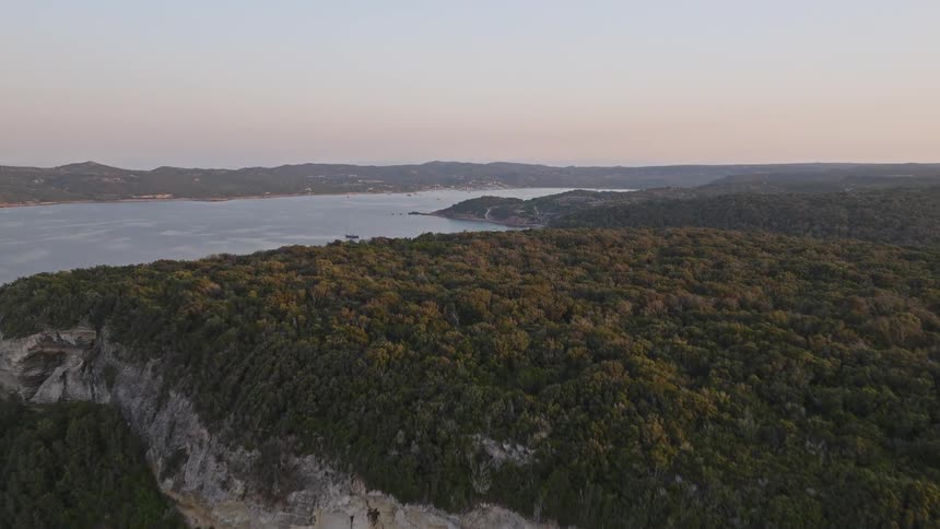 An aerial view rises over catamarans in a serene turquoise bay to reveal the stunning cliffs and lush green landscape of a coastal island at dusk