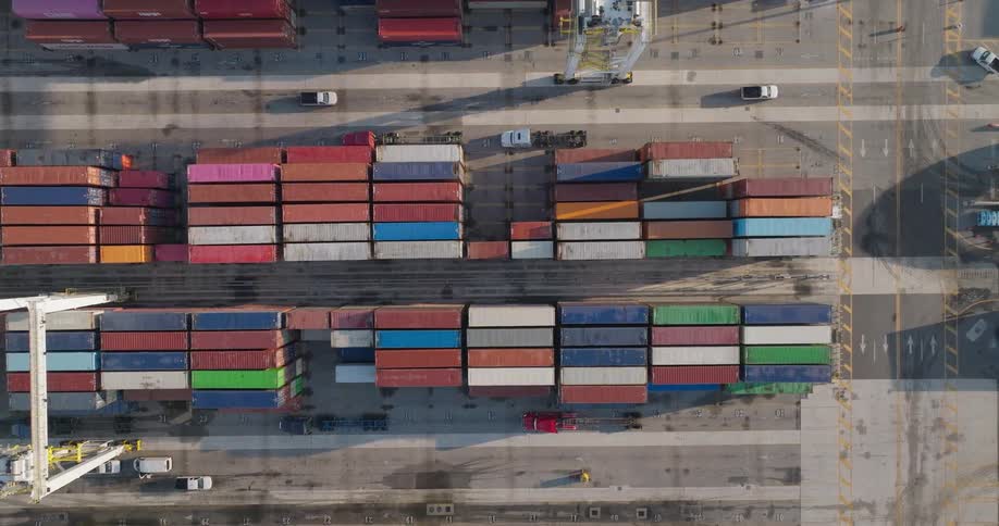 Aerial top-down shot of a busy shipping container terminal, with gantry cranes and trucks moving cargo through the industrial port