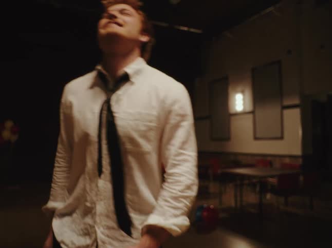 A joyful young man dances with wild abandon in a dimly lit hall before realizing he is all alone after the party
