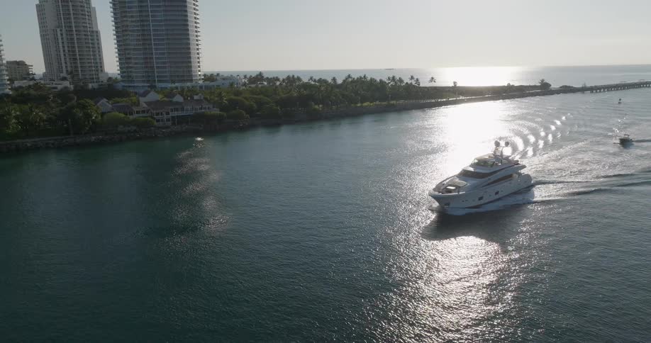 A stunning aerial view of a luxury yacht cruising along the coastline next to modern high-rise buildings as the sun glints on the water