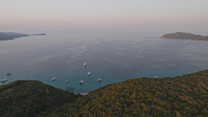 A serene aerial view shows yachts and catamarans anchored in a calm turquoise bay surrounded by lush green hills at sunset