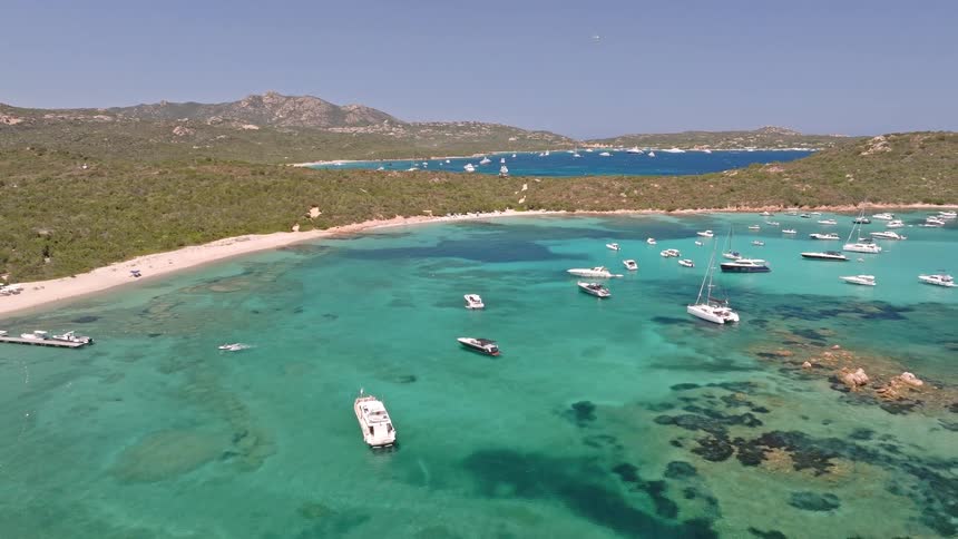An aerial view pulls back from a coastal pier to reveal a stunning turquoise bay filled with yachts and boats on a sunny summer day