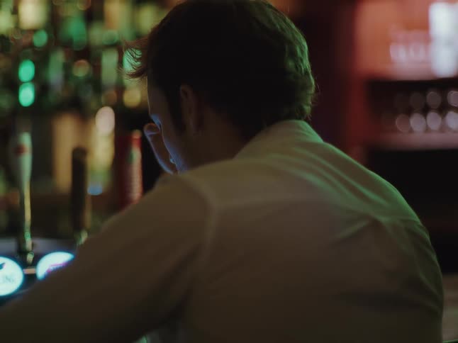 Over the shoulder shot of a man scrolling on his phone while sitting alone at a moody, dark bar