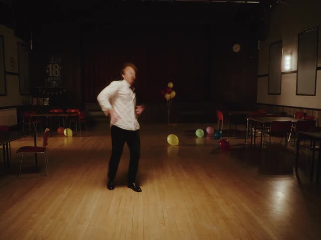 A man with ginger hair dances joyfully and wildly alone in an empty hall after his 18th birthday party celebrations