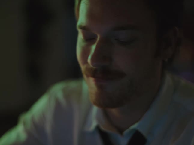 A stressed and contemplative man with a mustache closes his eyes and rubs his forehead, feeling overwhelmed in a dimly lit room at night