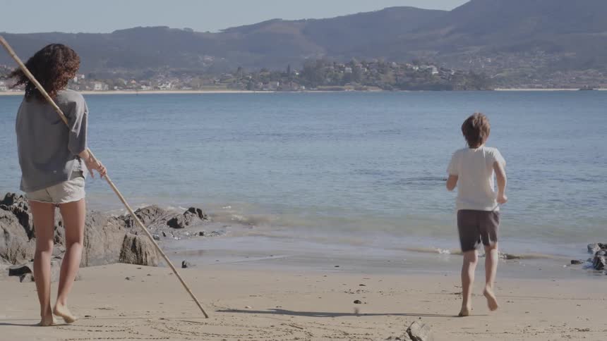 A young boy and a girl with curly hair play with long sticks on a sunny beach, running towards the ocean waves in slow motion
