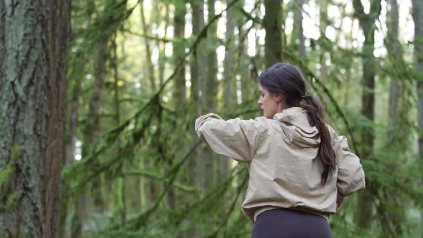 A woman runner in a beige jacket stretches and checks her smartwatch before continuing her run through a mossy, green forest