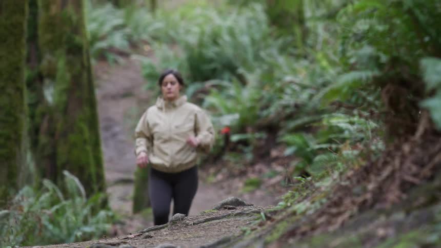 A woman in athletic wear jogs up a trail surrounded by mossy trees and ferns in slow motion