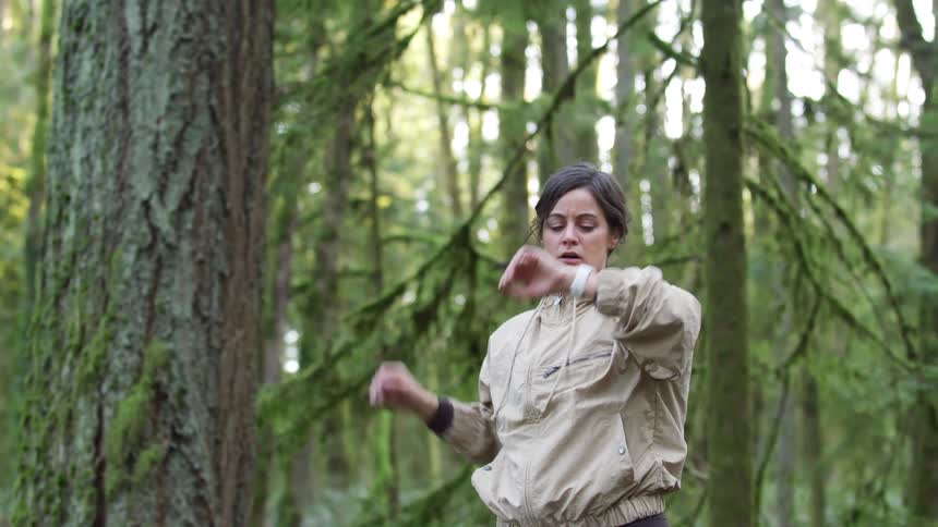 A woman trail running through a lush forest stops to catch her breath and checks her smartwatch before continuing on her run