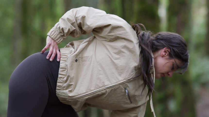 A fit woman in athletic clothing performs a deep stretch while warming up in a lush green forest