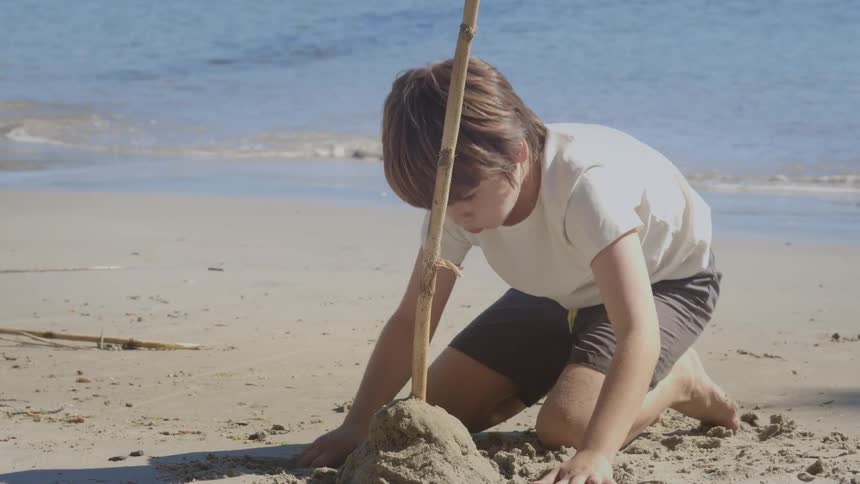 Focused boy builds a sand tower with a stick on the beach by the ocean waves
