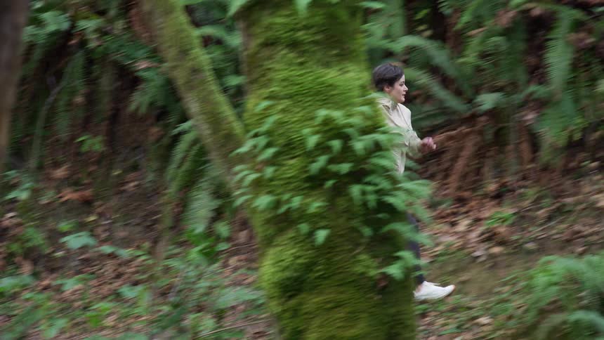 Side view of a female athlete trail running through a dense, mossy forest in slow motion