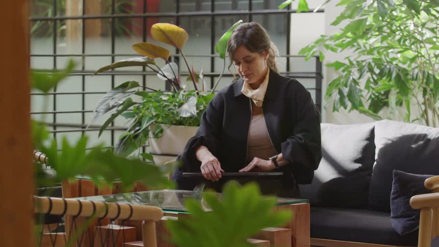 A woman gets ready to work on her laptop in a bright, modern patio surrounded by plants