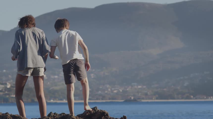 A boy and girl stand together on rocks by the sea, looking down as they enjoy the coastal mountain view