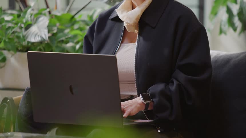 A woman works on her laptop while sitting on a couch in a modern office space surrounded by plants