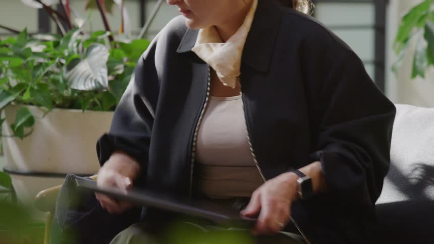 A woman finishes typing on her laptop, closes it, and packs it away in a plant-filled, sunlit modern room