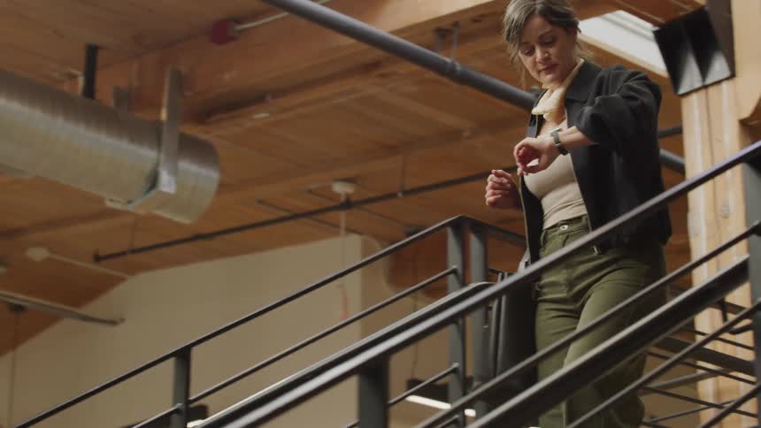 A stylish businesswoman walks up the stairs in a modern office, checking her smartwatch as she goes