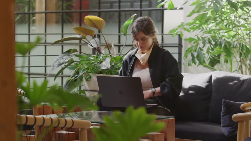 A woman works on her laptop in a sunlit, modern office lounge, then packs up her bag and leaves