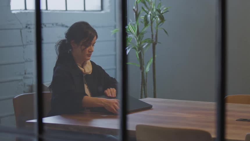 Viewed through a glass paneled window, a professional woman enters a conference room, sits down, and starts working on her laptop