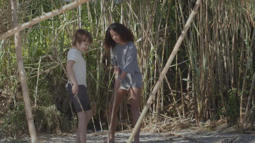 A young boy and girl have fun playing with sticks in the sand on a beach next to tall reeds
