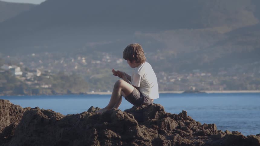 A young boy sits peacefully on a coastal rock, engrossed in play with the ocean and distant town in the background